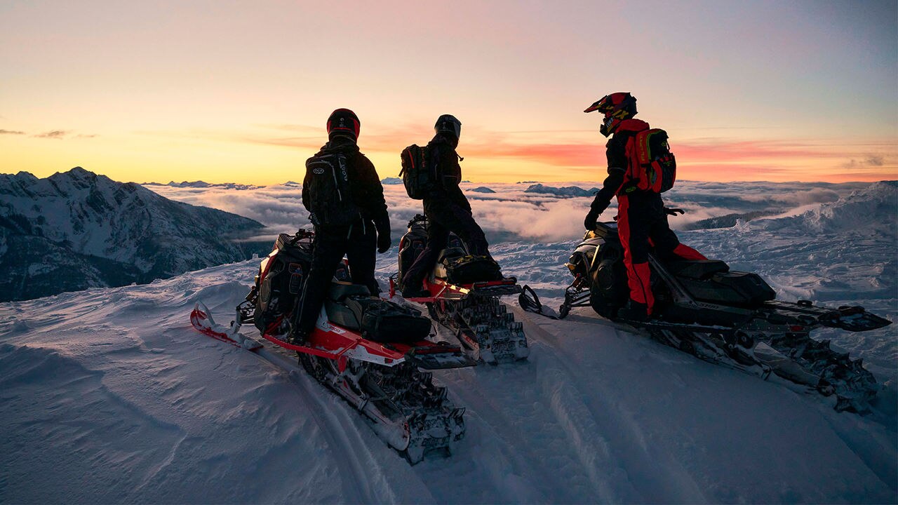 Tre førere på et fjell med Lynx-snøscootere når solen går ned.