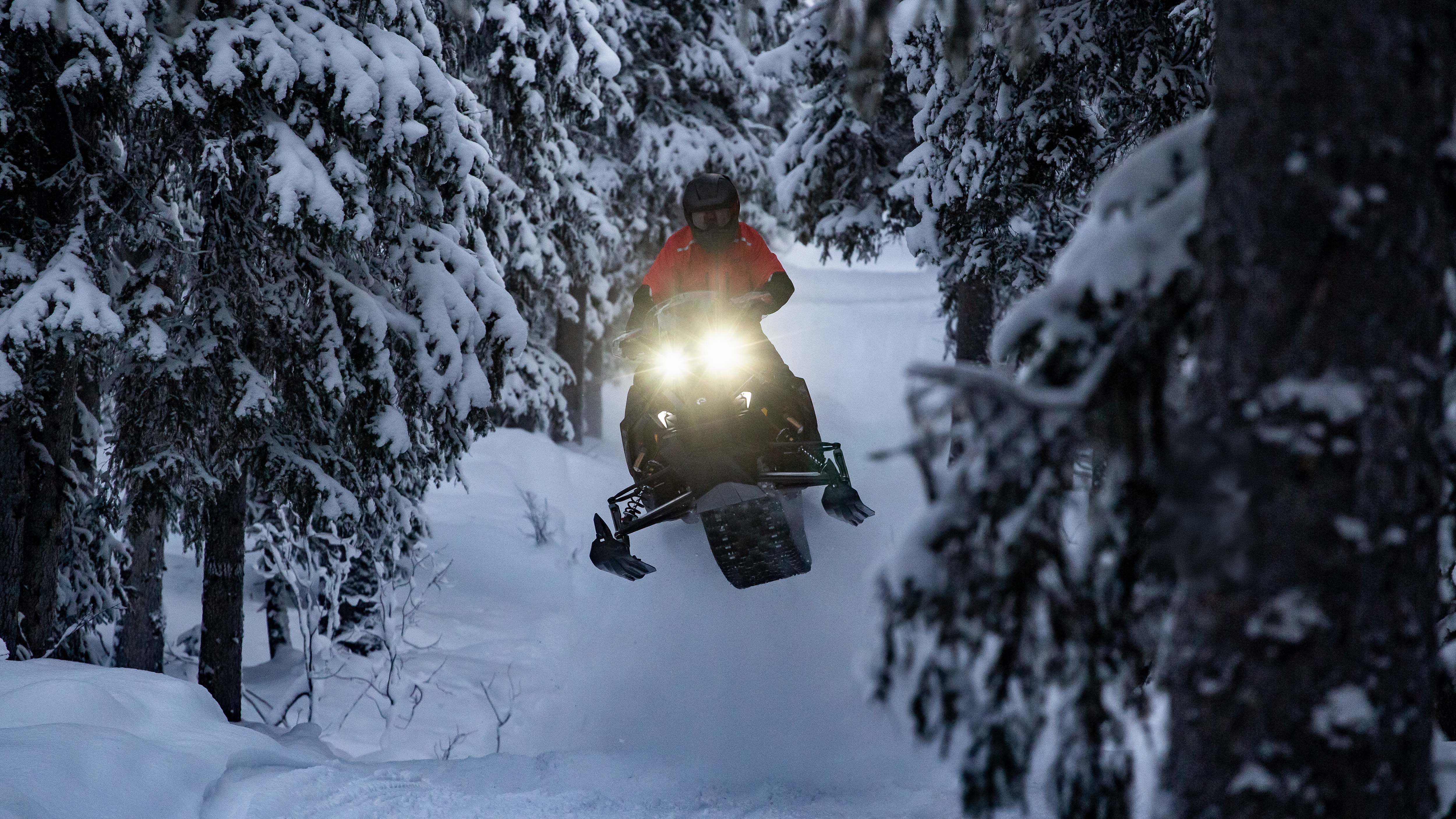 Rider on his 2026 Lynx Rave snowmobile in mid-air, jumping through a snowy forest trail with headlights shining bright.