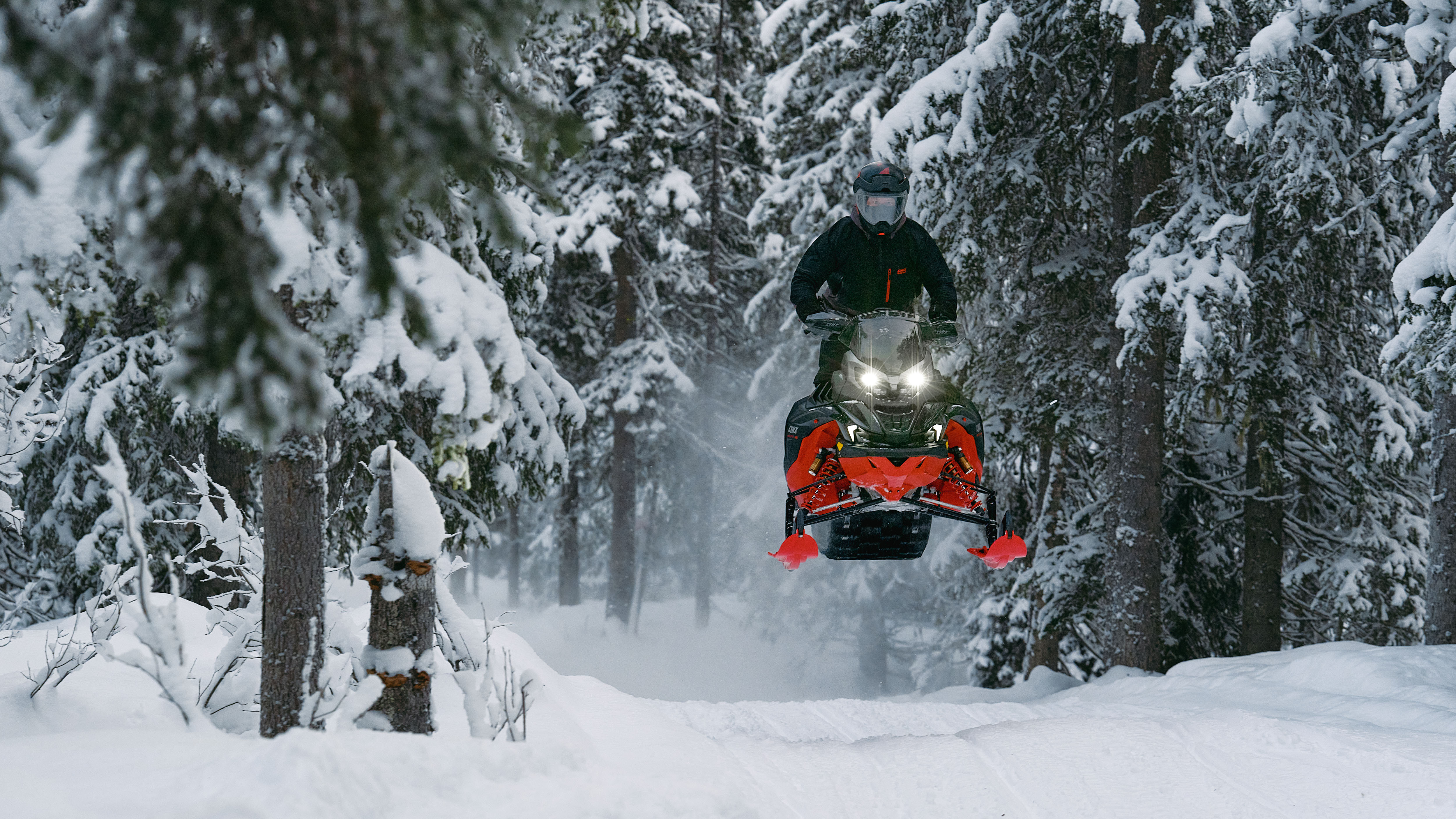 A snowmobiler on the 2026 Lynx Brutal RE snowmobile in mid-air in a snowy forest.