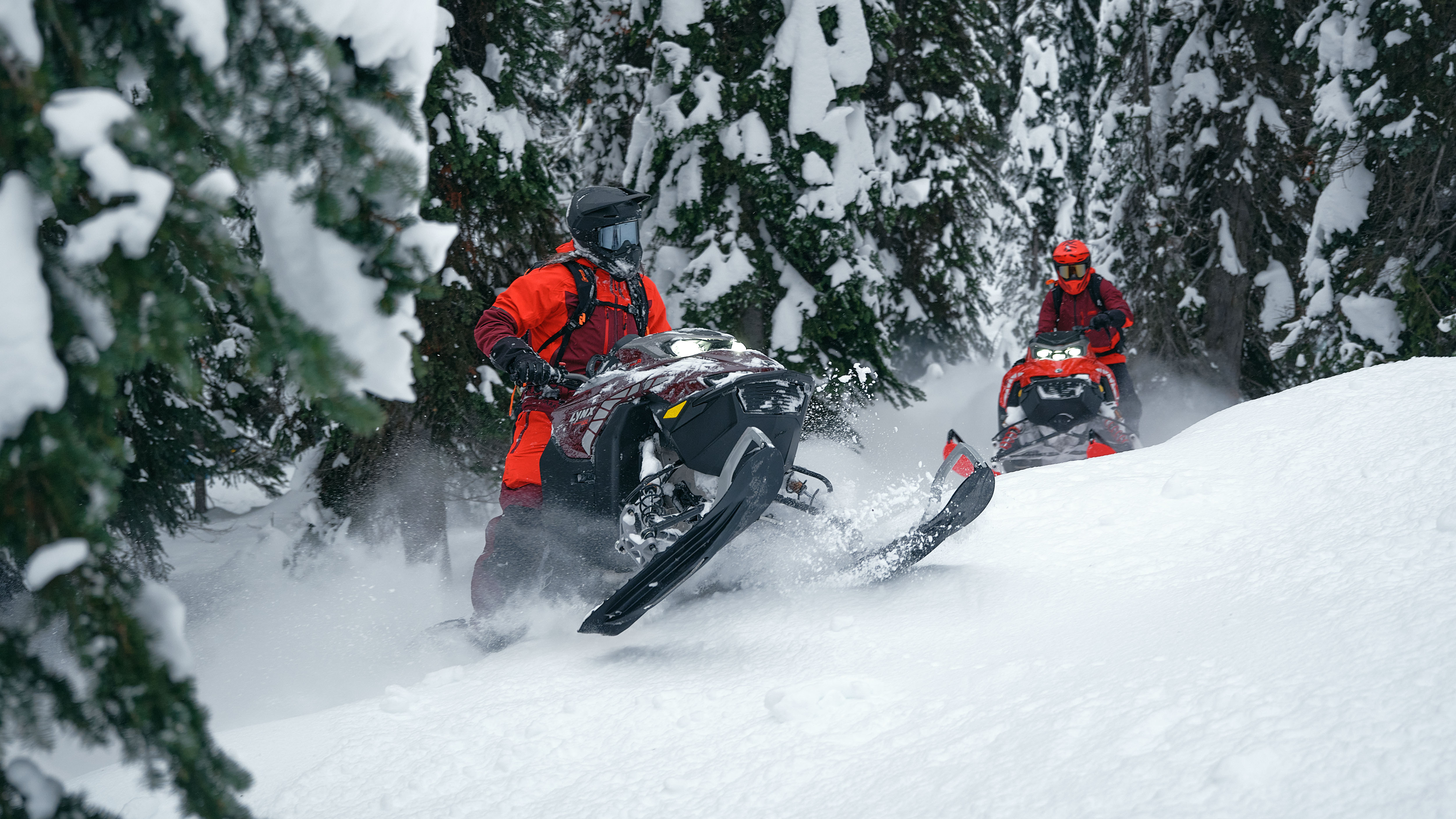 Two snowmobilers riding their 2026 Lynx Shredder snowmobile for deep snow in a snowy forest.