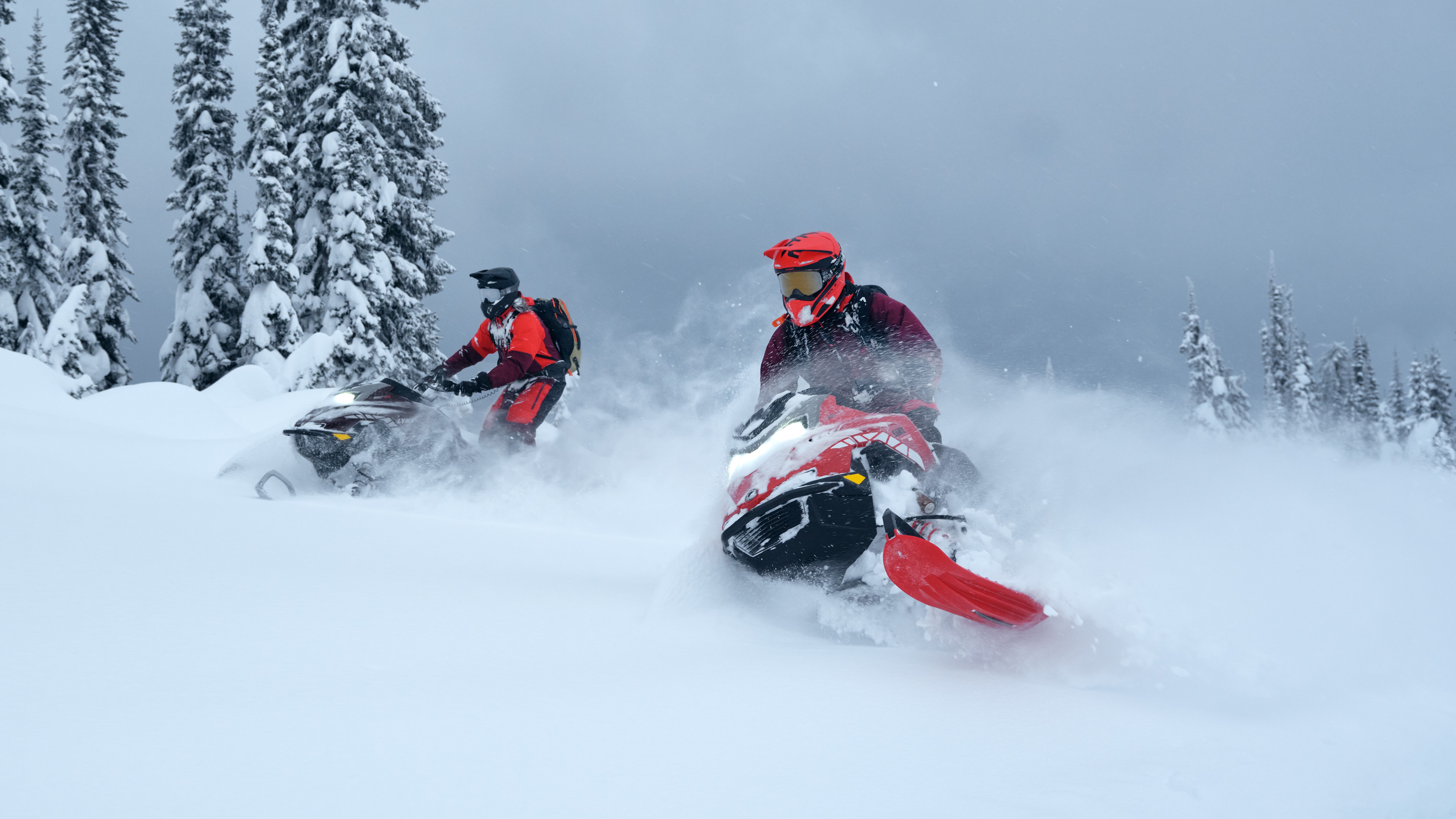 Two riders on their 2026 Lynx Shredder snowmobile carving through the fresh powder of a snow-covered forest.