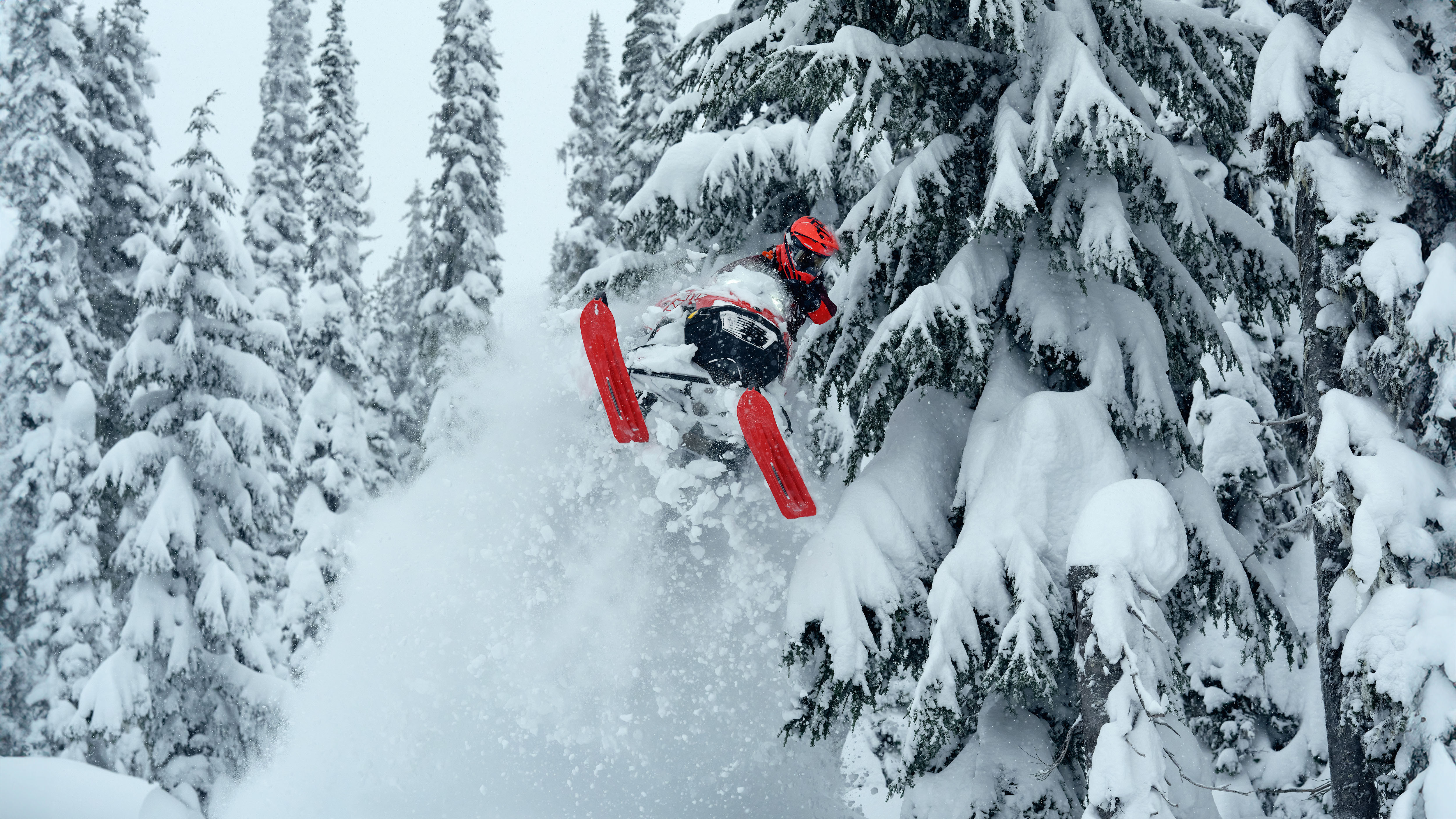 2026 Lynx Shredder RE snowmobile launches into a mid-air jump against a backdrop of a snowy, forested mountain landscape.