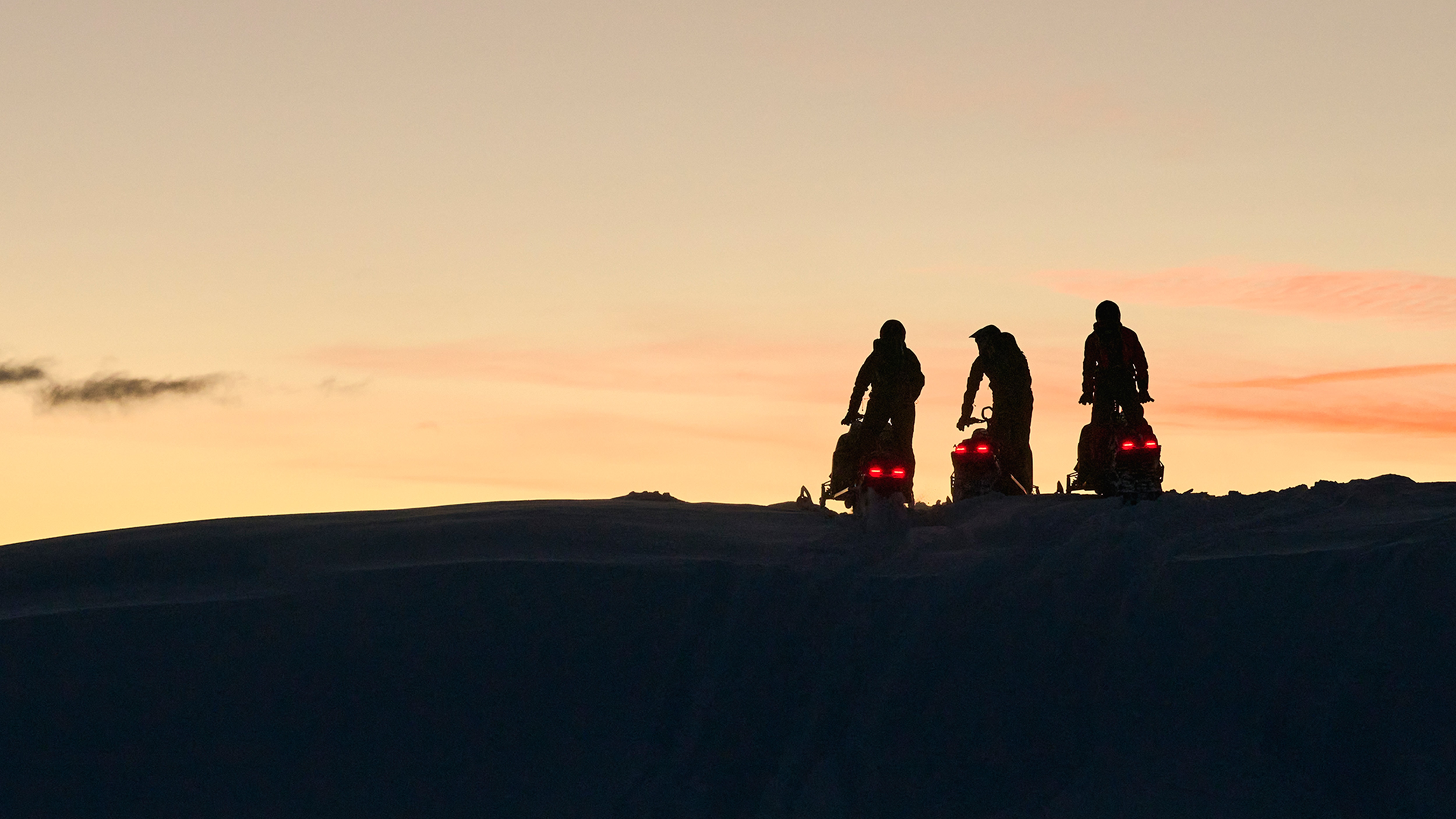 Tre snøscootere parkert på toppen av bakken ved solnedgang